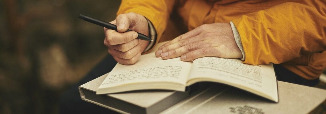 A person writing in a book with a pen and phone.